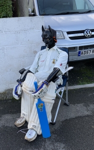 A devilish scarecrow wearing cricket pads and holding a bat!